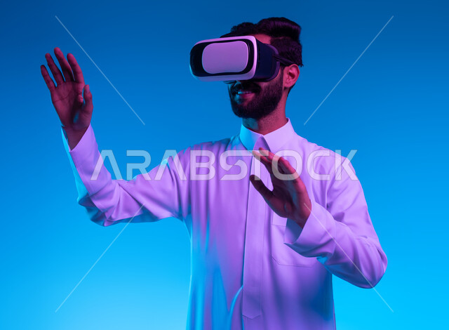 التفاعل ومحاكاة العالم الخيالي, بورتريه مقرب لشاب عربي خليجي سعودي يرتدي الثوب التقليدي و نظارات الواقع الافتراضي بايماءات الدفاع عن النفس، الاندماج ومحاولة امساك ولمس شيء ما, تقنيات حديثة وأجهزة تكنولوجية، خلفية زرقاء