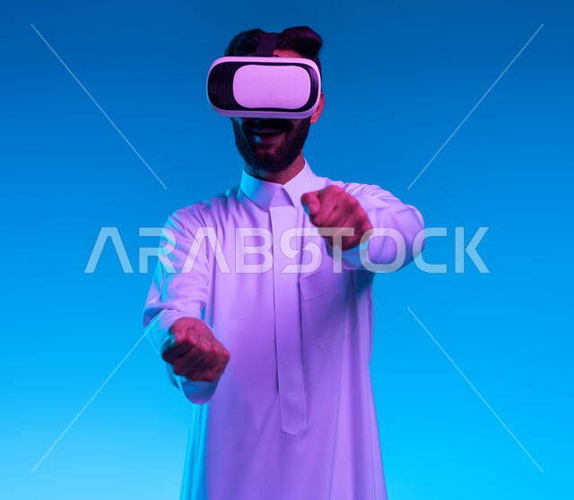 محاكاة ركوب سيارة ثري دي في العالم الخيالي، بورتريه مقرب لشاب عربي خليجي سعودي يرتدي الثوب التقليدي ونظارة الواقع الإفتراضي ثلاثية الأبعاد يقوم باللعب في ألعاب الفيديو، تقنيات وأجهزة تكنولوجية، إيماءات يدين تدل على السعادة بألعاب قيادة الدراجات، خلفية زرقاء