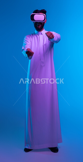 إيماءات يدين تدل على قيادة السيارة، أجهزة وتقنيات تكنولوجية، محاكاة ركوب سيارات ثري دي في العالم الخيالي، بورتريه لشاب عربي خليجي سعودي يرتدي الثوب التقليدي ونظارة الواقع الإفتراضي ثلاثية الأبعاد يقوم باللعب في ألعاب الفيديو، صورة الجسم بالكامل، خلفية زرقاء