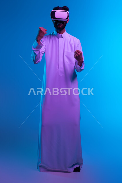 ممارسة رياضة الملاكمة والمصارعة بتقنيات تكنولوجية، تفريغ الطاقة السلبية، التدرب على الحركات القتالية من خلال ألعاب الفيديو، بورتريه لشاب عربي خليجي سعودي يرتدي الثوب التقليدي ونظارات الواقع الافتراضي بإيماءات الدفاع عن النفس، صورة الجسم بالكامل، خلفية زرقاء