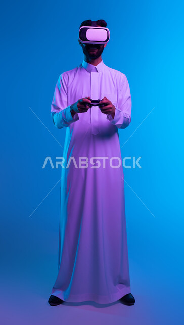 تقنيات حديثة وأجهزة تكنولوجية، بورتريه لشاب عربي خليجي سعودي يرتدي الثوب التقليدي ونظارة الواقع الإفتراضي يقف بإستقامة يمسك بيده عصا التحكم يقوم باللعب في ألعاب الفيديو الترفيهية، دمج العالم الخيالي مع المتعة والتسلية، صورة الجسم بالكامل، خلفية زرقاء