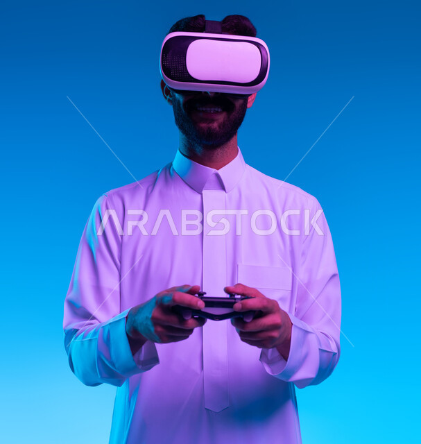 دمج العالم الخيالي مع المتعة والتسلية، بورتريه مقرب لشاب عربي خليجي سعودي مبتسم يرتدي الثوب التقليدي ونظارة الواقع الإفتراضي يقف بإستقامة يمسك بيده عصا التحكم يقوم باللعب في ألعاب الفيديو الترفيهية، أجهزة تكنولوجية وتقنيات حديثة، خلفية زرقاء
