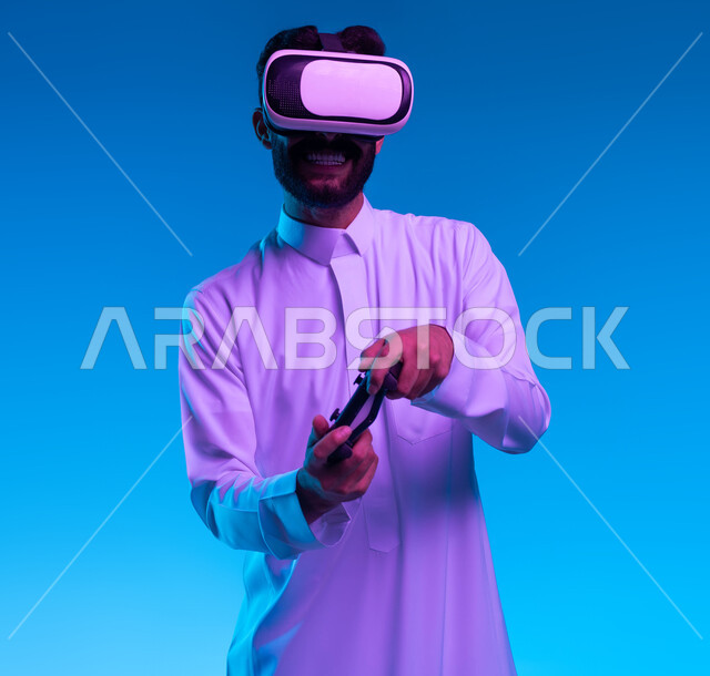 دمج المتعة والتسلية مع العالم الخيالي، بورتريه مقرب لشاب عربي خليجي سعودي مبتسم يرتدي الثوب التقليدي ونظارة الواقع الإفتراضي يمسك بيده عصا التحكم يقوم باللعب في ألعاب الفيديو الترفيهية، أجهزة تكنولوجية وتقنيات حديثة، قضاء اوقات فراغ ممتعة، خلفية زرقاء