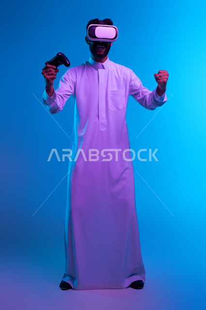 ايماءات تدل على الانتصار والفوز، دمج المتعة والتسلية مع العالم الخيالي، بورتريه لشاب عربي خليجي سعودي يرتدي الثوب التقليدي ونظارة الواقع الإفتراضي يمسك بيده عصا التحكم يلعب في ألعاب الفيديو الترفيهية، أجهزة تكنولوجية حديثة، صورة الجسم بالكامل، خلفية زرقاء