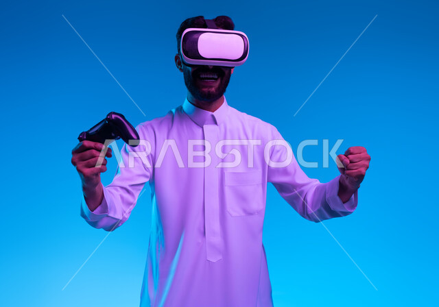 تعابير تدل على الانتصار والفوز، دمج المتعة والتسلية مع العالم الخيالي، بورتريه مقرب لشاب عربي خليجي سعودي مبتسم يرتدي الثوب التقليدي ونظارة الواقع الإفتراضي يمسك بيده عصا التحكم يقوم باللعب في ألعاب الفيديو الترفيهية، أجهزة تكنولوجية وتقنيات حديثة، خلفية زرقاء