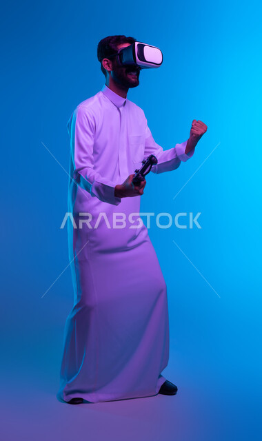 ايماءات تدل على الانتصار والفوز، دمج المتعة والتسلية مع العالم الخيالي، بورتريه لشاب عربي خليجي سعودي يرتدي الثوب التقليدي ونظارة الواقع الإفتراضي يمسك بيده عصا التحكم يلعب في ألعاب الفيديو الترفيهية، أجهزة تكنولوجية حديثة، صورة الجسم بالكامل، خلفية زرقاء