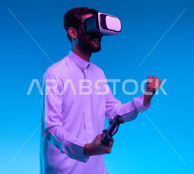 تعابير تدل على الانتصار والفوز، دمج المتعة والتسلية مع العالم الخيالي، بورتريه مقرب لشاب عربي خليجي سعودي مبتسم يرتدي الثوب التقليدي ونظارة الواقع الإفتراضي يمسك بيده عصا التحكم يقوم باللعب في ألعاب الفيديو الترفيهية، أجهزة تكنولوجية وتقنيات حديثة، خلفية زرقاء