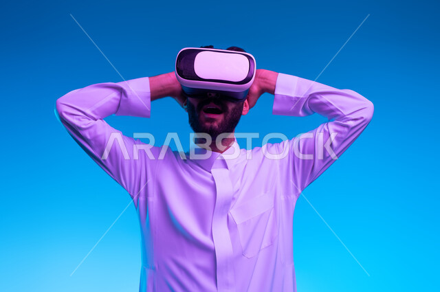 ايماءات وجه ويدين تدل على الدهشة والاستغراب, مشاهدة الأفلام عبر نظارات في آر، بورتريه مقرب لرجل عربي خليجي سعودي يرتدي الثوب التقليدي ونظارة الواقع الافتراضي ثلاثية الأبعاد يقوم بالتفاعل مع الفلم ومحاكاة العالم الخيالي، خلفيه زرقاء