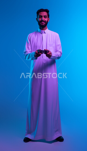 دمج المتعة والتسلية مع العالم الالكتروني، بورتريه لشاب عربي خليجي سعودي مبتسم يرتدي الثوب التقليدي يقف باستقامة يمسك بيده عصا التحكم يقوم باللعب في ألعاب الفيديو الترفيهيه، تعابير تدل على الحماس والإنسجام، أجهزة تكنولوجية حديثة، صورة الجسم بالكامل، خلفية زرقاء