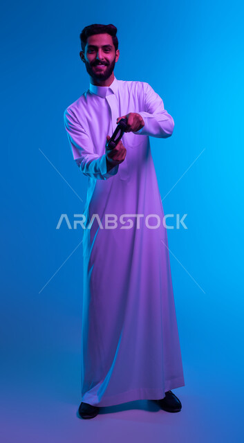 النظر الى الكاميرا بتعابير تدل على الحماس والإنسجام، دمج المتعة والتسلية مع العالم الالكتروني، بورتريه لشاب عربي خليجي سعودي مبتسم يرتدي الثوب التقليدي يمسك بيده عصا التحكم يقوم باللعب في ألعاب الفيديو، أجهزة تكنولوجية حديثة، صورة الجسم بالكامل، خلفية زرقاء