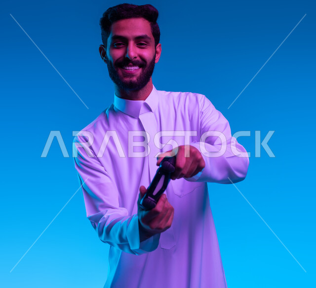 النظر الى الكاميرا بتعابير تدل على الحماس والإنسجام، دمج المتعة والتسلية مع العالم الالكتروني، بورتريه مقرب لشاب عربي خليجي سعودي مبتسم يرتدي الثوب التقليدي يمسك بيده عصا التحكم يقوم باللعب في ألعاب الفيديو الترفيهيه، أجهزة تكنولوجية وتقنيات حديثة، خلفية زرقاء