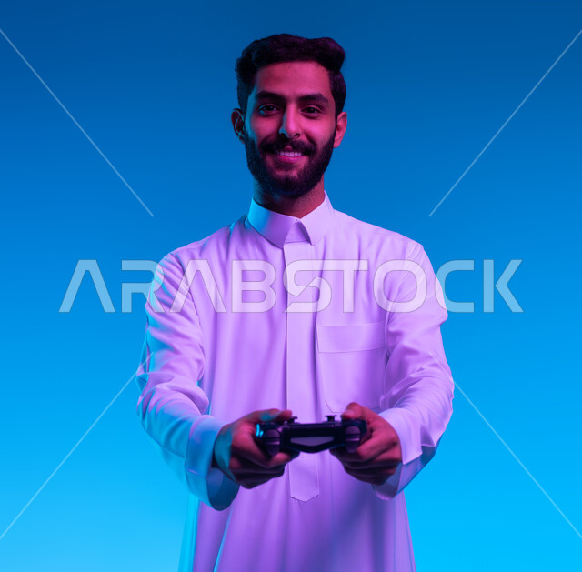 دمج المتعة والتسلية مع العالم الالكتروني، بورتريه مقرب لشاب عربي خليجي سعودي مبتسم يرتدي الثوب التقليدي يمسك بيده عصا التحكم يقوم باللعب في ألعاب الفيديو الترفيهيه، النظر الى الكاميرا بتعابير تدل على الحماس والإنسجام، أجهزة تكنولوجية وتقنيات حديثة، خلفية زرقاء