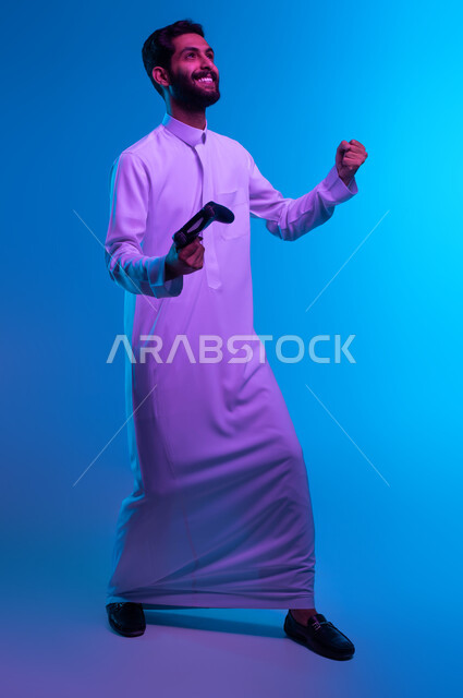 إيماءات تدل على الانتصار والفوز، دمج العالم الالكتروني مع التسلية والمتعة، بورتريه لشاب عربي خليجي سعودي متحمس يرتدي الثوب التقليدي يمسك بيده عصا التحكم يقوم باللعب في ألعاب الفيديو الترفيهية، أجهزة تكنولوجية حديثة، صورة الجسم بالكامل، خلفية زرقاء