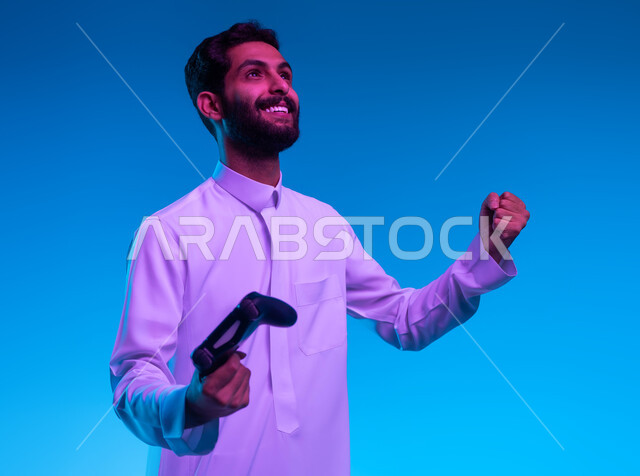 تعابير تدل على الانتصار والفوز، دمج العالم الالكتروني مع التسلية، بورتريه مقرب لشاب عربي خليجي سعودي مبتسم يرتدي الثوب التقليدي يمسك بيده عصا التحكم يقوم باللعب في ألعاب الفيديو الترفيهية، أجهزة تكنولوجية حديثة، خلفية زرقاء