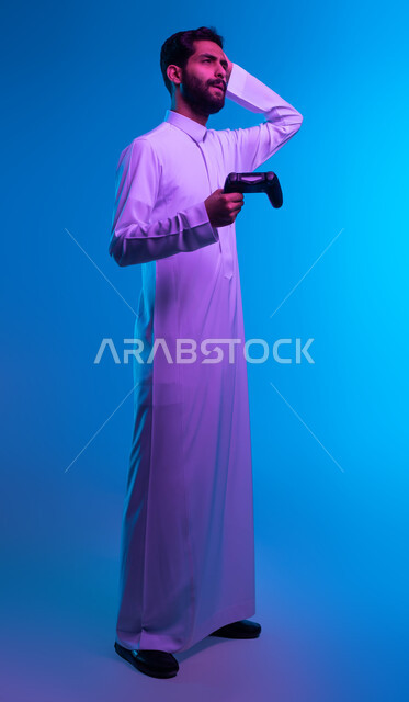 إيماءات الخسارة وخيبة الأمل، التسلية والترفيه بالفيديو جيمز في وقت الفراغ، استخدام الألعاب الالكترونية والأجهزة الحديثة، بورتريه لشاب عربي خليجي سعودي يرتدي الثوب التقليدي يمسك عصا التحكم ويضع يده على رأسه بحسرة، صورة الجسم بالكامل، خلفية زرقاء