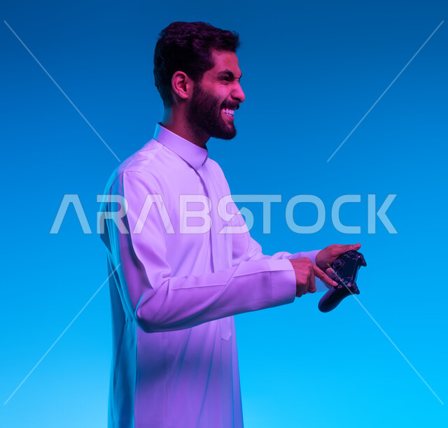 العالم الرقمي الترفيهي، تجربة ترفيهية ممتعة ومثيرة في الألعاب الالكترونية والأجهزة الحديثة، المتعة والتسلية بالفيديو جيمز، بورتريه مقرب لشاب عربي خليجي سعودي يرتدي الثوب التقليدي يمسك بيده عصا التحكم بإيماءات التفاعل والاندماج والسعادة، خلفية زرقاء