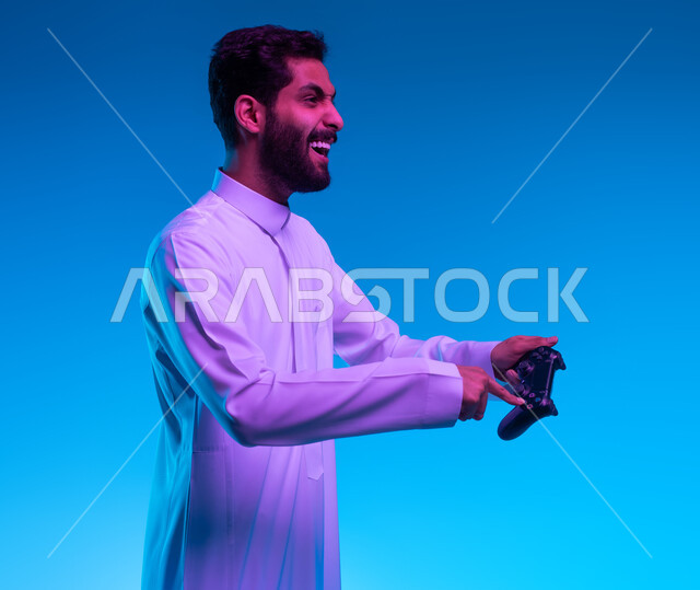 العالم الرقمي الترفيهي، تجربة ترفيهية ممتعة ومثيرة في الألعاب الالكترونية والأجهزة الحديثة، المتعة والتسلية بالفيديو جيمز، بورتريه مقرب لشاب عربي خليجي سعودي يرتدي الثوب التقليدي يمسك بيده عصا التحكم بإيماءات التفاعل والاندماج والسعادة، خلفية زرقاء