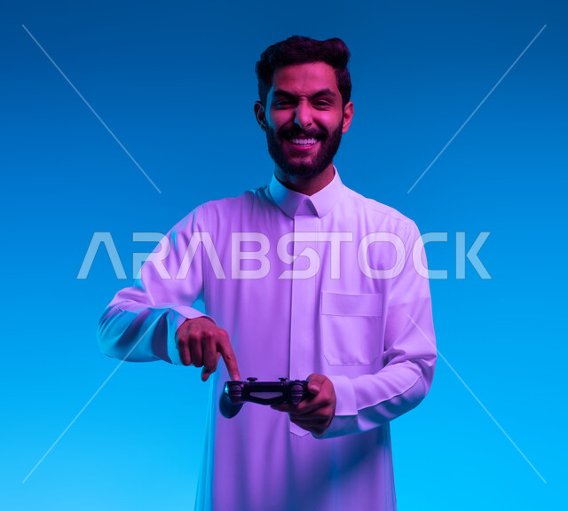 المتعة والتسلية بالفيديو جيمز، العالم الرقمي الترفيهي، تجربة ترفيهية ممتعة ومثيرة في الألعاب الالكترونية والأجهزة الحديثة، بورتريه مقرب لشاب عربي خليجي سعودي يرتدي الثوب التقليدي يمسك بيده عصا التحكم بإيماءات التفاعل والاندماج والسعادة، خلفية زرقاء