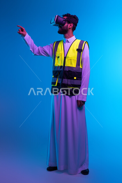 تطور ونمو المجال الهندسي عبر نظارة الفي آر، رؤية مستقبلية سعودية 2030، التفاعل مع الهندسة الافتراضية، بورتريه لمهندس عربي خليجي سعودي يرتدي السترة الخاصة بالعمل ونظارة الواقع الافتراضي يشير إلى عنصر ما، صورة لكامل الجسم، خلفية زرقاء