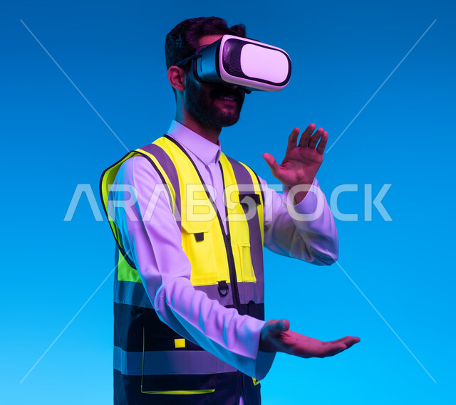 التفاعل مع الهندسة الافتراضية، بورتريه مقرب لمهندس عربي خليجي سعودي يرتدي السترة الخاصة بالعمل ونظارة الواقع الافتراضي يقوم بحمل عنصر ما، رؤية مستقبلية 2030، نمو وتطور المجال الهندسي عبر نظارة الفي آر، استخدام التكنولوجيا في المجالات الهندسية، خلفية زرقاء