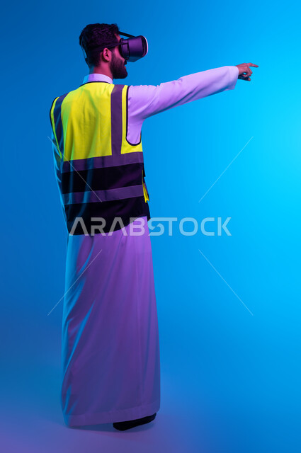 التفاعل  والاندماج مع الهندسة الافتراضية، تطور المجالات الهندسية عبر نظارات الفي آر، رؤية مستقبلية سعودية 2030، بورتريه من الخلف لمهندس عربي خليجي سعودي يرتدي السترة الخاصة بالعمل ونظارة الواقع الافتراضي يشير إلى عنصر ما، صورة لكامل الجسم، خلفية زرقاء