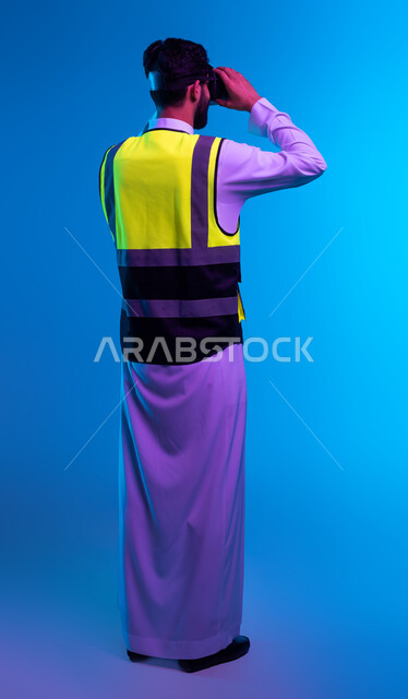 Technological progress and development in architecture, future vision 2030, simulation of imaginary reality, interaction with virtual engineering fields, portrait from behind of a Saudi Gulf Arab engineer wearing a work jacket and VR glasses, full body portrait, blue background