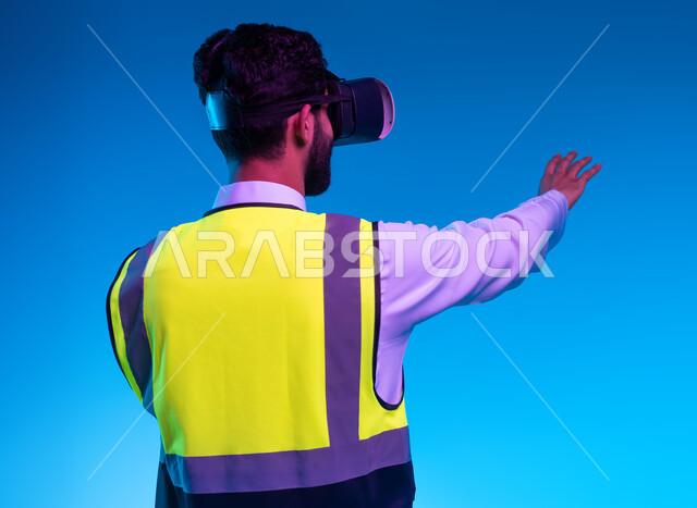 The use of technology in engineering fields, interaction with virtual engineering, the development of the engineering field through VR glasses, future vision 2030, a close-up portrait from the back of a Saudi Gulf Arab engineer wearing his work jacket and virtual reality glasses trying to hold an item, blue background
