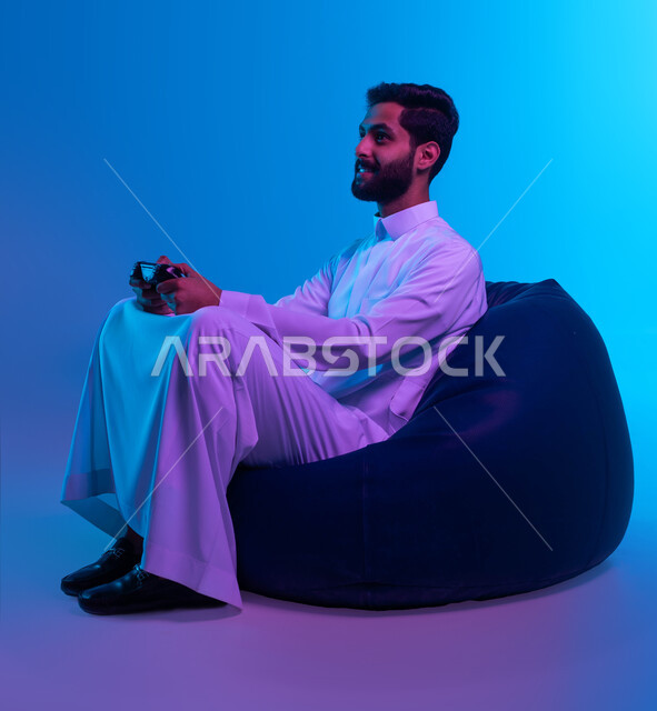 تجربة ترفيهية ممتعة ومثيرة في الألعاب الالكترونية والأجهزة الحديثة، العالم الرقمي الترفيهي، المتعة والتسلية بالفيديو جيمز، بورتريه لشاب عربي خليجي سعودي يرتدي الثوب التقليدي يمسك بيده عصا التحكم بإيماءات التفاعل والاندماج، خلفية زرقاء