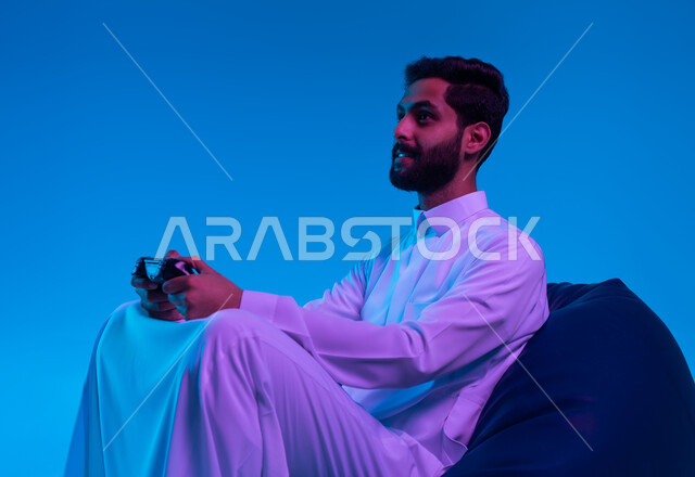 العالم الرقمي الترفيهي، تجربة ترفيهية ممتعة ومثيرة في الألعاب الالكترونية والأجهزة الحديثة، المتعة والتسلية بالفيديو جيمز، بورتريه مقرب لشاب عربي خليجي سعودي يرتدي الثوب التقليدي يمسك بيده عصا التحكم بإيماءات التفاعل والاندماج، خلفية زرقاء