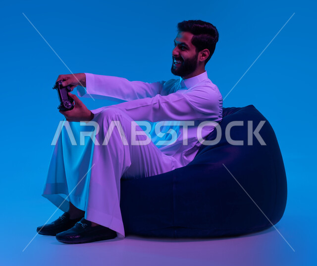 الاستمتاع  باللعب بالألعاب الالكترونية الترفيهية، استخدام الأجهزة التكنولوجية والتقنيات الحديثة، بورتريه لشاب عربي خليجي سعودي مبتسم يرتدي الثوب التقليدي يمسك بيده عصا التحكم بإيماءات التفاعل والاندماج، خلفية زرقاء