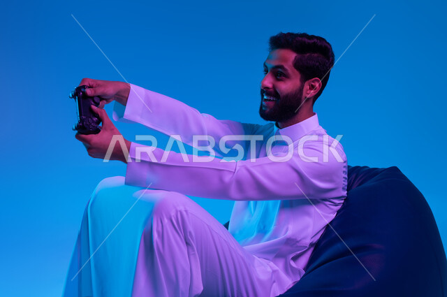 استخدام الأجهزة التكنولوجية والتقنيات الحديثة، الاستمتاع  باللعب بالألعاب الالكترونية الترفيهية، بورتريه لشاب عربي خليجي سعودي مبتسم يرتدي الثوب التقليدي يمسك بيده عصا التحكم بإيماءات التفاعل والاندماج، خلفية زرقاء