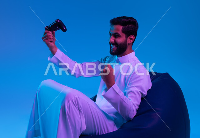 التفاعل والاندماج أثناء اللعب بالألعاب الالكترونية الترفيهية، المتعة والتسلية بالفيديو جيمز، بورتريه لشاب عربي خليجي سعودي مبتسم يرتدي الثوب التقليدي يمسك بيده عصا التحكم ويرفع قبضته للأعلى بإيماءات الفوز والانتصار، خلفية زرقاء