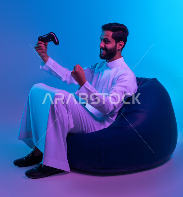 المتعة والتسلية بالفيديو جيمز، التفاعل والاندماج أثناء اللعب بالألعاب الالكترونية الترفيهية، بورتريه لشاب عربي خليجي سعودي مبتسم يرتدي الثوب التقليدي يمسك بيده عصا التحكم ويرفع قبضته للأعلى بإيماءات الفوز والانتصار، خلفية زرقاء