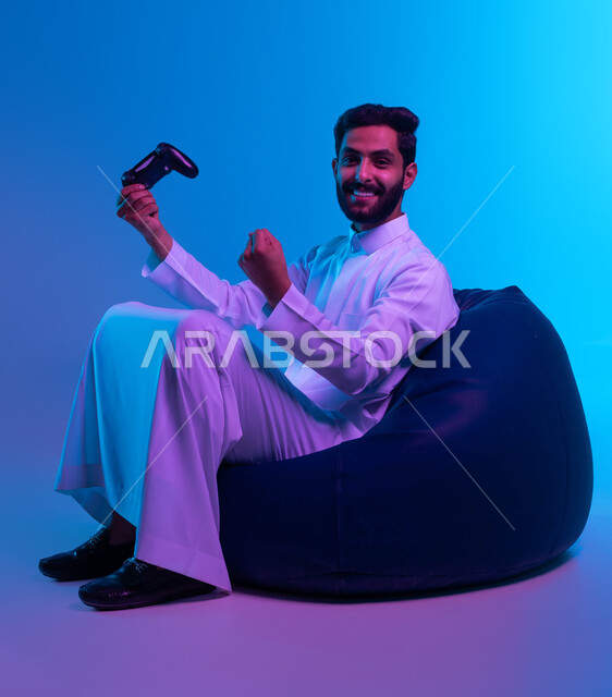 Interaction and integration while playing entertainment electronic games, fun and entertainment in video games, a portrait of a smiling Gulf Arab Saudi man wearing a traditional dress holding a joystick in his hand and raising his fist upwards with gestures of victory and victory, blue background