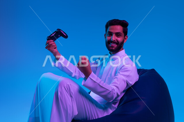 المتعة والتسلية بالفيديو جيمز، التفاعل والاندماج أثناء اللعب بالألعاب الالكترونية الترفيهية، بورتريه لشاب عربي خليجي سعودي مبتسم يرتدي الثوب التقليدي يمسك بيده عصا التحكم ويرفع قبضته للأعلى بإيماءات الفوز والانتصار، خلفية زرقاء