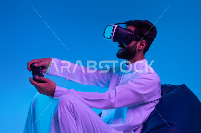 استخدام الأجهزة التكنولوجية والتقنيات الحديثة، دمج المتعة والتسلية مع الواقع الافتراضي والخيالي، بورتريه مقرب لشاب عربي خليجي سعودي مبتسم يرتدي الثوب التقليدي ونظارة الفي آر يمسك بيده عصا التحكم يقوم باللعب في ألعاب الفيديو الترفيهية، خلفية زرقاء