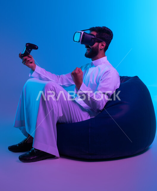 العالم الترفيهي الرقمي، تجربة مثيرة في الألعاب الالكترونية، التسلية في أوقات الفراغ بالفيديو جيمز، بورتريه لشاب عربي خليجي سعودي مسرور يرتدي الثوب التقليدي ونظارة في آر يجلس على كرسي بين باج و يمسك يد التحكم ويرفع قبضته بإيماءات النصر، خلفية زرقاء