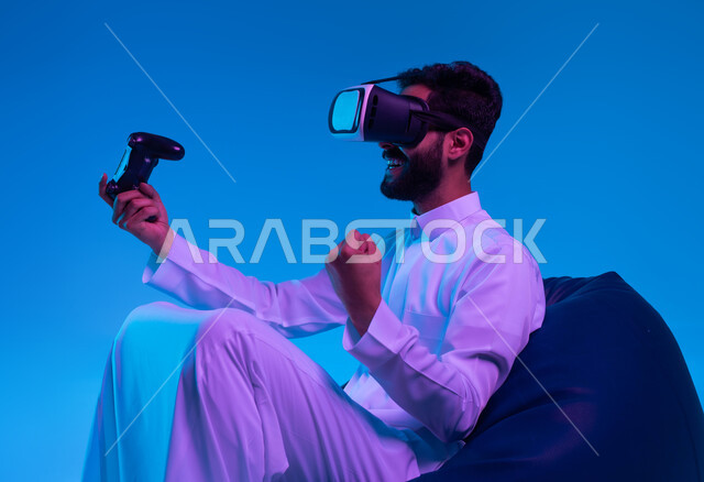 تجربة مثيرة في الألعاب الالكترونية، التسلية في أوقات الفراغ بالفيديو جيمز، العالم الرقمي الترفيهي، بورتريه لشاب عربي خليجي سعودي مسرور يرتدي الثوب التقليدي ونظارة في آر يجلس على كرسي بين باج و يمسك يد التحكم ويرفع قبضته بإيماءات النصر، خلفية زرقاء