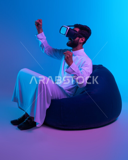 الشغف في متابعه فريق كرة القدم، ايماءات الحماس والاندماج ,التفاعل ومحاكاة العالم الخيالي, رفع اليدين عاليات بحركات الفوز، بورتريه لشاب عربي خليجي سعودي يرتدي الثوب التقليدي يجلس على كرسي بين باج و يقوم بمشاهدة أفلام عبر نظارة الواقع الافتراضي, خلفية زرقاء