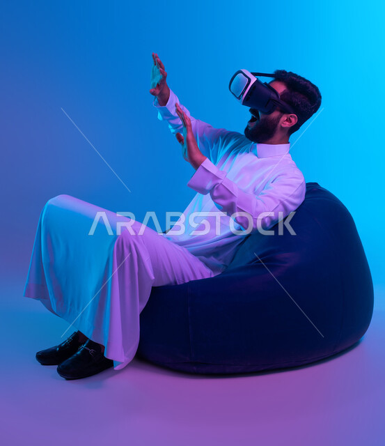 Gestures of enthusiasm and integration, interaction and simulation of the imaginary world, entertaining modern techniques, trying to hold something with movements of amazement, a portrait of a Saudi Gulf Arab young man wearing a traditional dress, sitting on a bean bag chair and watching movies through virtual reality glasses, blue background.