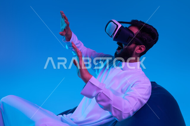 Trying to catch something with movements of amazement, interaction and simulation of the imaginary world, gestures of enthusiasm and integration, modern entertaining techniques, a close-up portrait of a Saudi Gulf Arab young man wearing a traditional thobe, sitting on a bean bag chair and watching movies through virtual reality glasses, blue background.