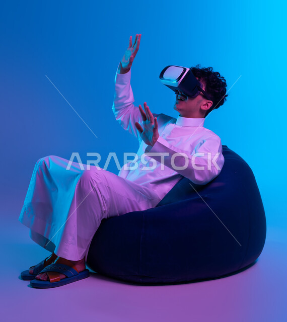 Interaction and simulation of the imaginary world, enthusiasm, integration, and trying to catch something, modern entertaining techniques, a portrait of a Saudi Gulf Arab young man wearing a traditional dress, sitting on a bean bag chair and watching movies through 3D virtual reality glasses, blue background.