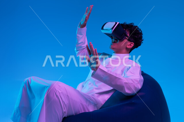 Enthusiasm, integration, and trying to catch something, modern entertaining techniques, interaction and simulation of the imaginary world, portrait of a Saudi Gulf Arab young man wearing a traditional thobe, sitting on a bean bag chair and watching movies through 3D virtual reality glasses, blue background.