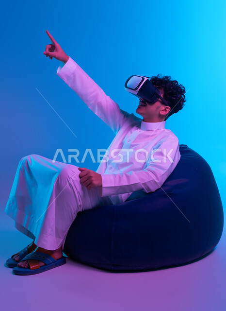 Simulating an imaginary world, using the sense of touch to touch an item with tap gestures, advanced modern technologies, a portrait of a Saudi Gulf Arab young man wearing a traditional thobe and 3D virtual reality glasses sitting on a bean bag chair and pointing at something with his index finger, blue background.