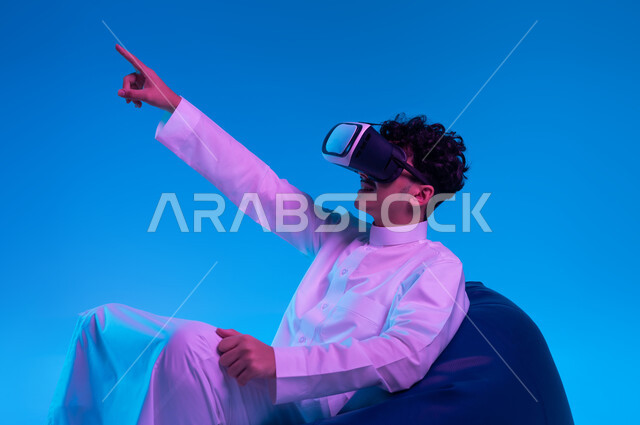 استخدام حاسة اللمس لملامسة عنصر ما بإيماءات النقر, تقنيات حديثة متطورة، بورتريه لشاب عربي خليجي سعودي يرتدي الثوب التقليدي ونظارة الواقع الافتراضي ثلاثية الأبعاد يجلس على كرسي بين باج و يشير إلى شيء ما بأصبع السبابة, محاكاة العالم الخيالي, خلفية زرقاء
