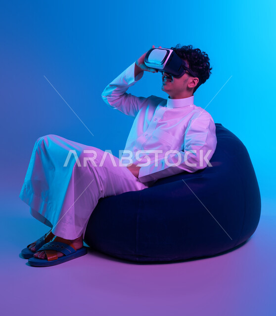 Using advanced modern technologies, interaction, integration, and simulation of the imaginary world, amusement, entertainment, and enjoyment in leisure time, a portrait of a Saudi Gulf Arab young man wearing traditional clothing and virtual reality glasses, sitting on a bean bag chair and watching favorite movies, blue background.