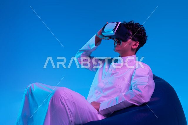 Interaction, integration and simulation of the imaginary world, amusement, entertainment and leisure time, use of advanced modern technologies, portrait of a Saudi Gulf Arab young man wearing traditional clothing and virtual reality glasses sitting on a bean bag chair and watching favorite movies, blue background