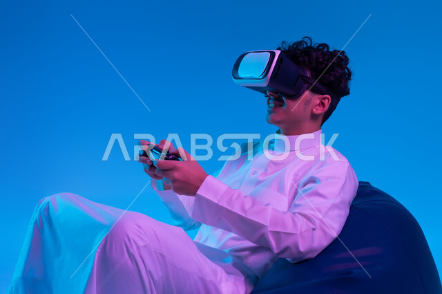 تقنيات حديثة مسلية, التفاعل والاندماج مع ألعاب الفيديو الترفيهية، استخدام الأجهزة التكنولوجية المتطورة، بورتريه لشاب عربي خليجي سعودي مبتسم يرتدي الثوب التقليدي ونظارة في آر يجلس على كرسي بين باج و يمسك يد التحكم ، خلفية زرقاء