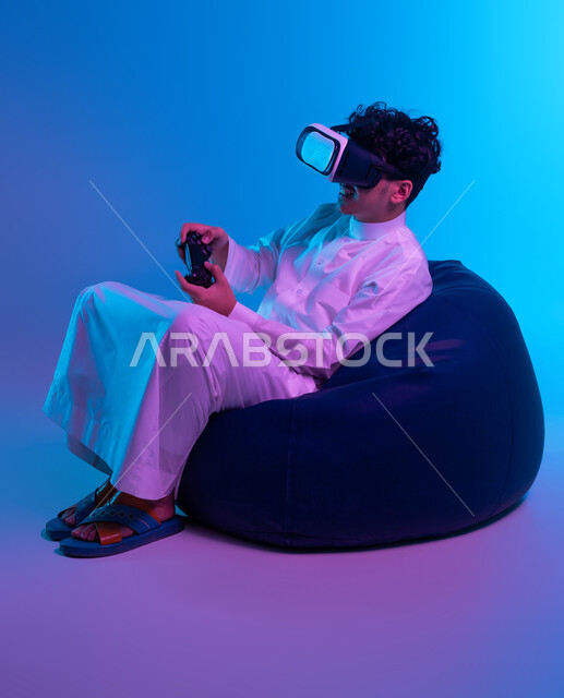 اللعب في ألعاب الفيديو الترفيهية، دمج التسلية مع الواقع الافتراضي والخيالي، استخدام الأجهزة التكنولوجية والتقنيات الحديثة، بورتريه لشاب عربي خليجي سعودي يرتدي الثوب التقليدي ونظارة في آر يجلس على كرسي بين باج و يمسك يد التحكم ، خلفية زرقاء