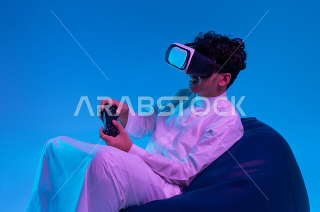 دمج التسلية مع الواقع الافتراضي والخيالي، استخدام الأجهزة التكنولوجية والتقنيات الحديثة، بورتريه لشاب عربي خليجي سعودي يرتدي الثوب التقليدي ونظارة في آر يجلس على كرسي بين باج و يمسك يد التحكم يقوم باللعب في ألعاب الفيديو الترفيهية، خلفية زرقاء